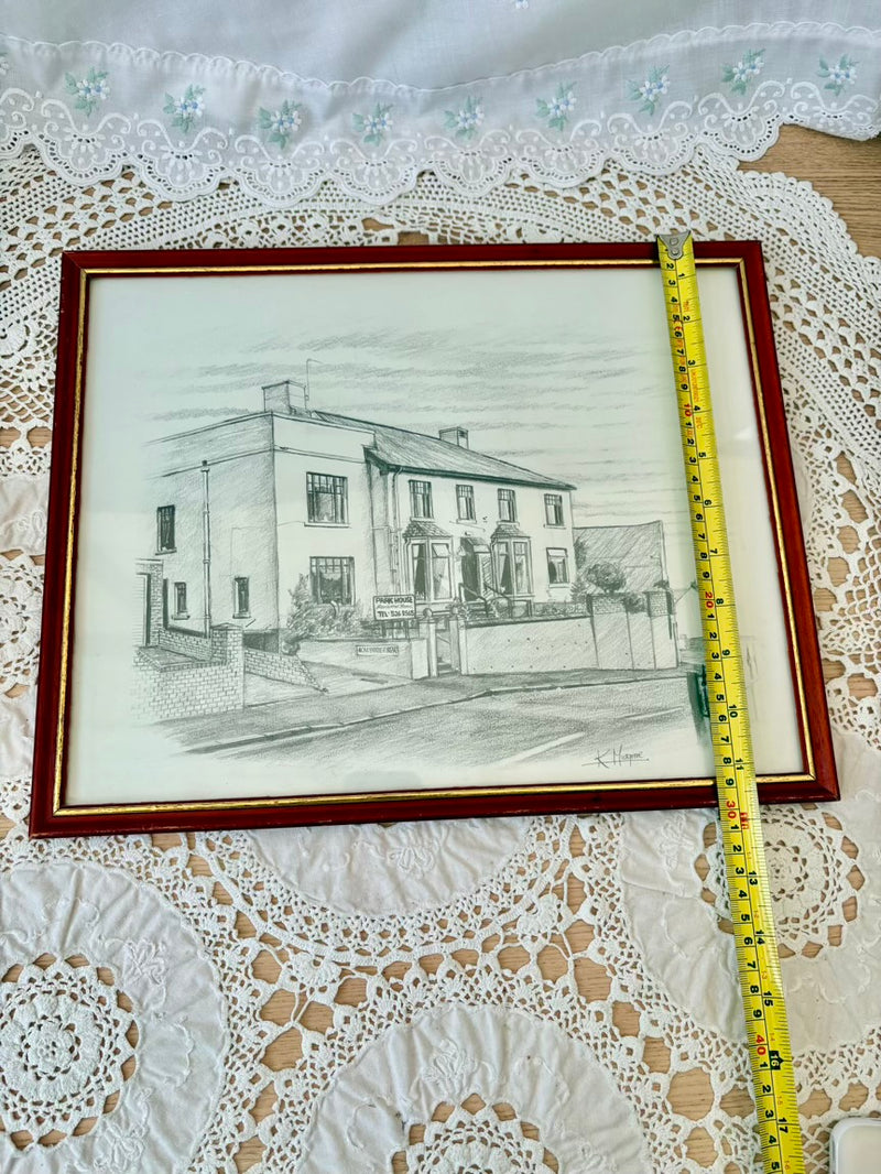 Framed pencil drawing of a house on a lace tablecloth with a measuring tape.