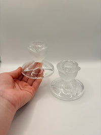 Clear glass candle holders on a white surface with a hand holding one of them.