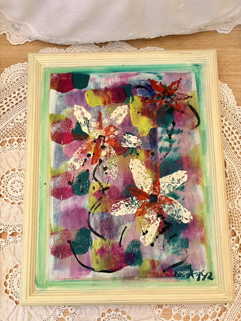 Colorful abstract painting of flowers in a light wooden frame on a lace tablecloth.