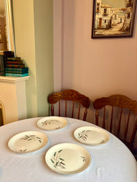 Dining table with four ceramic plates and two wooden chairs in a room with striped wallpaper and a framed picture on the wall.
