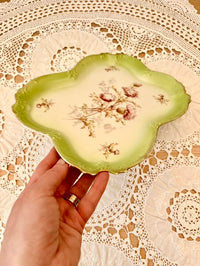 Hand holding a decorative green plate with floral patterns on a lace tablecloth.