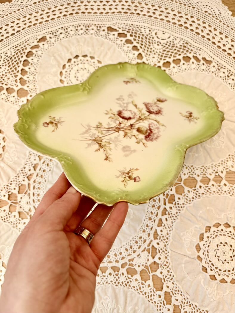 Hand holding a decorative green plate with floral patterns on a lace tablecloth.