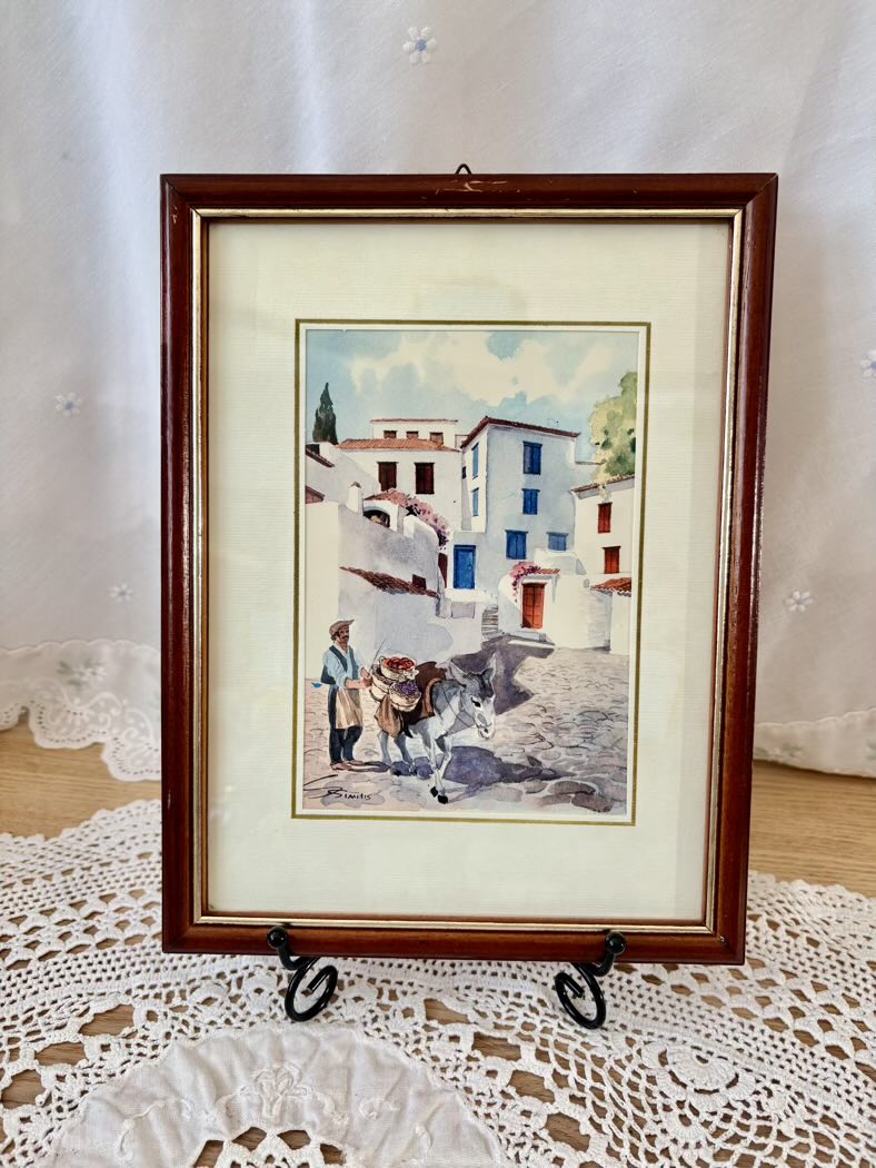 Framed artwork of a street scene with buildings and people on a lace tablecloth.