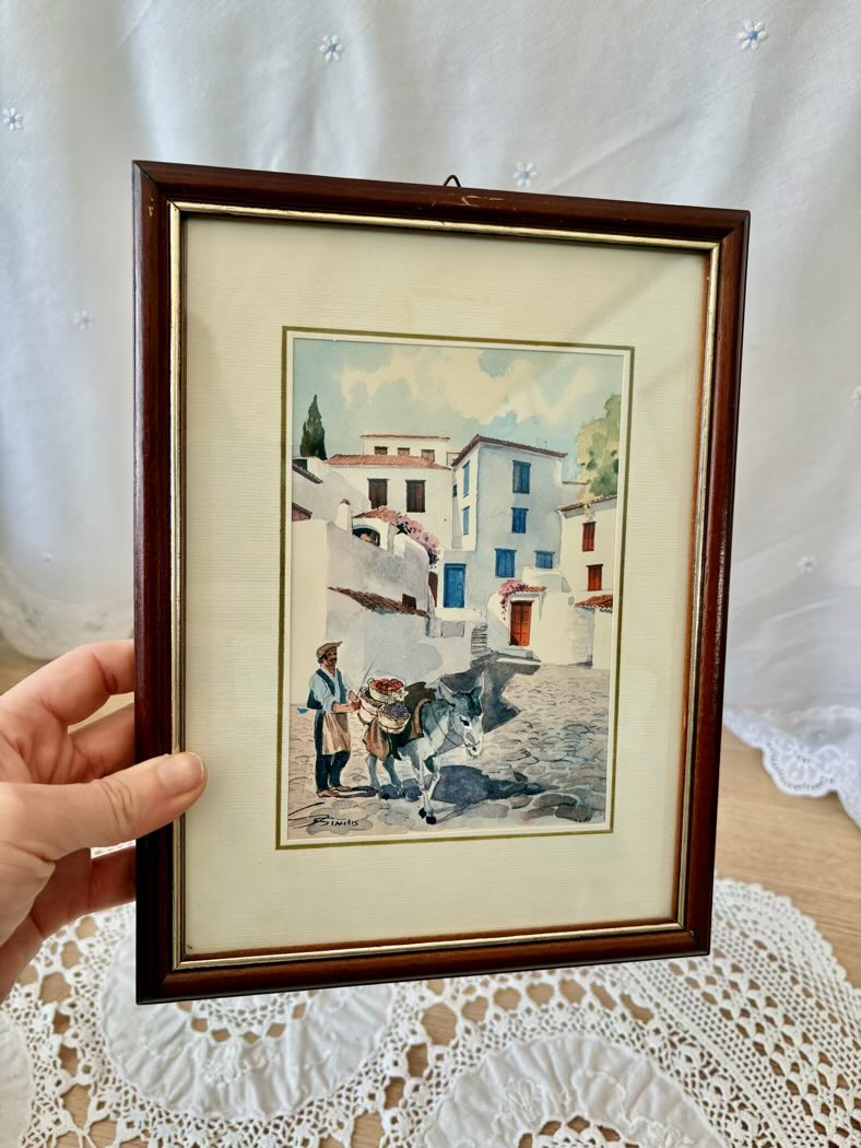 Framed artwork of a village scene with a person and horse on a lace tablecloth.