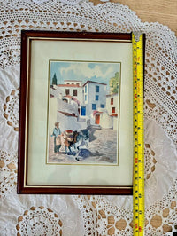 Framed artwork of a village scene with a cow, on a lace tablecloth with a tape measure for scale.