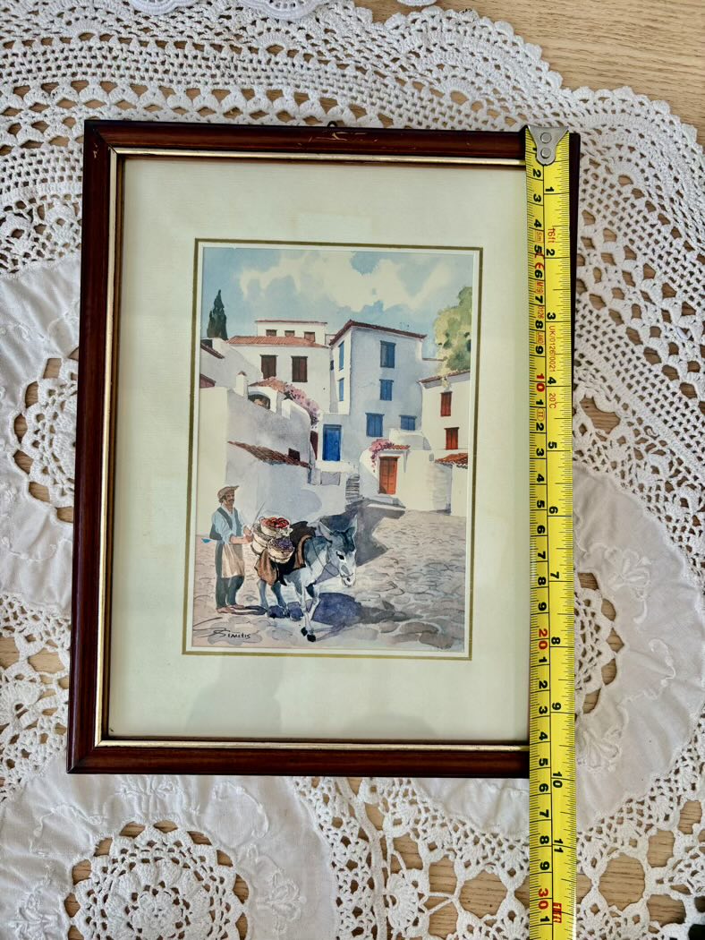 Framed artwork of a village scene with a cow, on a lace tablecloth with a tape measure for scale.