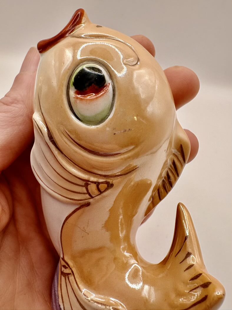 Ceramic fish-shaped object with a large eye held in a hand against a neutral background