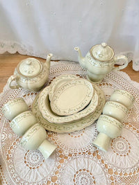 Ceramic tea set with teapot, cups, and plates on a lace tablecloth.