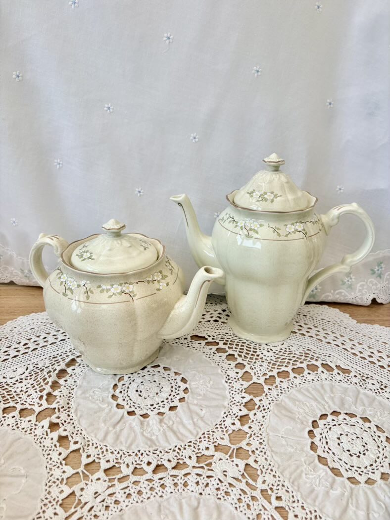 Two white ceramic teapots on a lace doily with a white curtain background