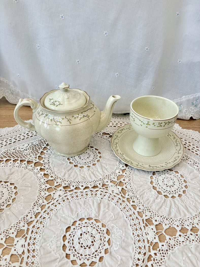 Teapot, cup, and saucer set on a lace tablecloth with a white curtain background
