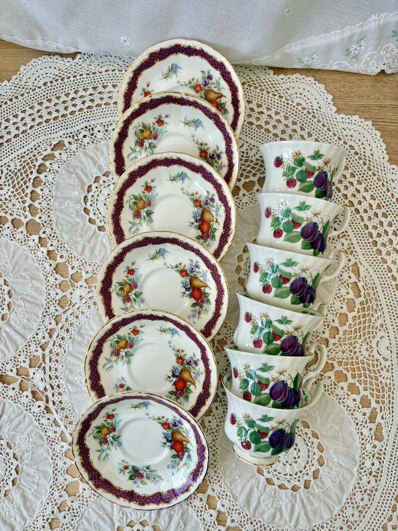 Set of ceramic teacups and saucers with floral and fruit patterns on a lace tablecloth.