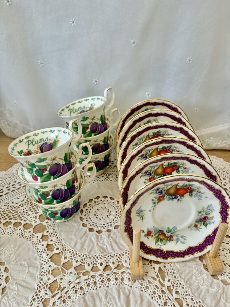 Set of ceramic teacups and saucers with fruit design on a lace tablecloth.