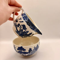 Ceramic pitcher with blue floral patterns being held over a matching bowl on a neutral background