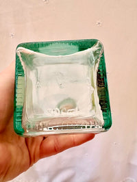 Clear glass square object with green rim held in a hand against a neutral background