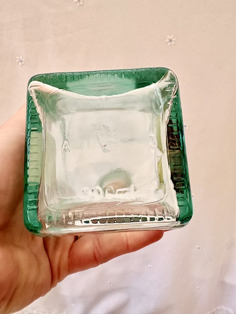 Clear glass square object with green rim held in a hand against a neutral background