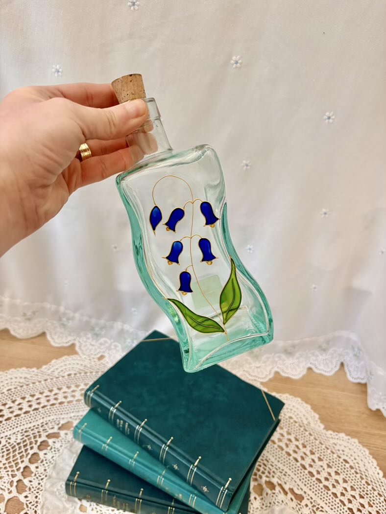 Hand holding a decorative glass bottle with blue flowers on a lace tablecloth.