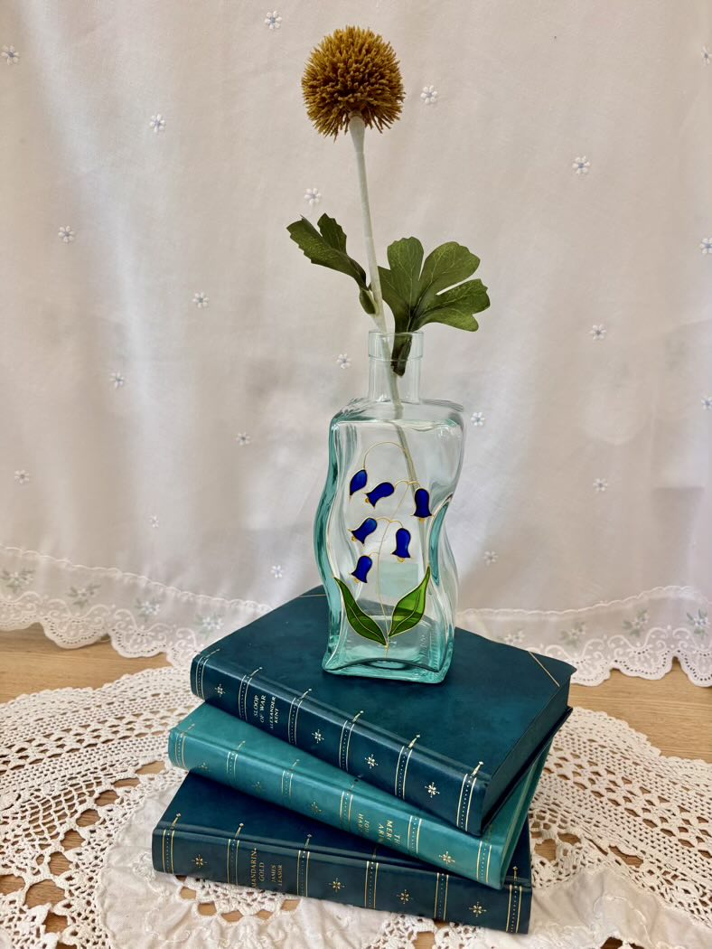 Decorative glass vase with flowers on top of stacked blue books against a lace-draped window.