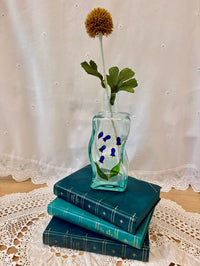 Decorative glass vase with flowers on top of stacked blue books against a lace-draped window.