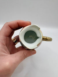 Hand holding a small ceramic mug with gold rim and handle against a plain background