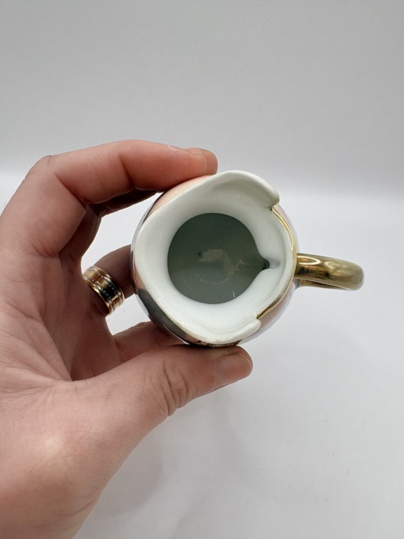 Hand holding a small ceramic mug with gold rim and handle against a plain background