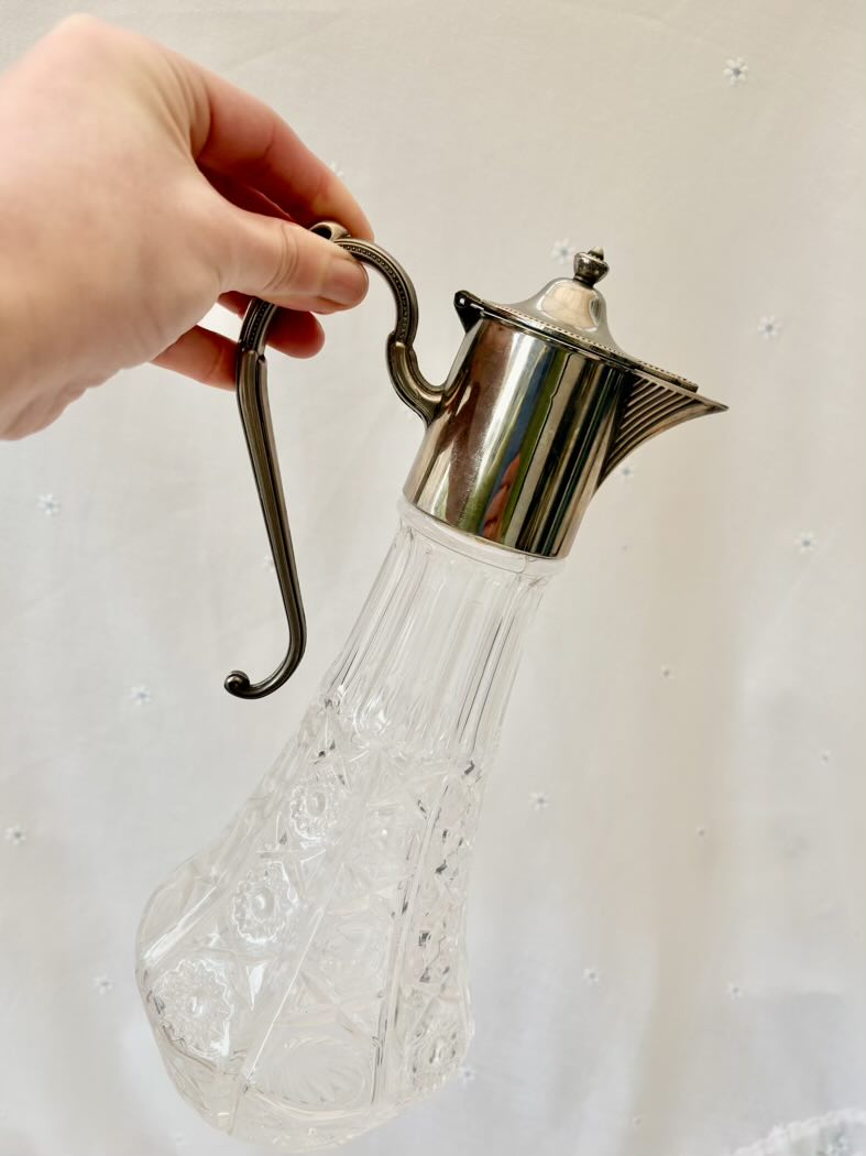 Clear glass carafe with silver metal top and handle held by a hand against a light background