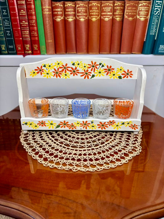 Decorative stand with colorful floral design holding small glass containers on a wooden surface.