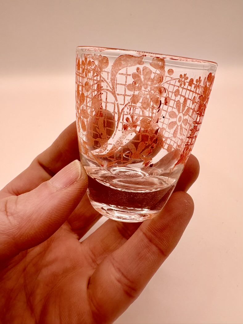 Hand holding a small glass with intricate red patterns on a plain background
