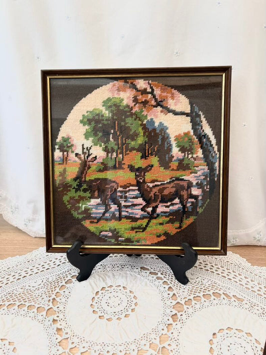 Framed needlepoint of two deer in a landscape on a white doily.