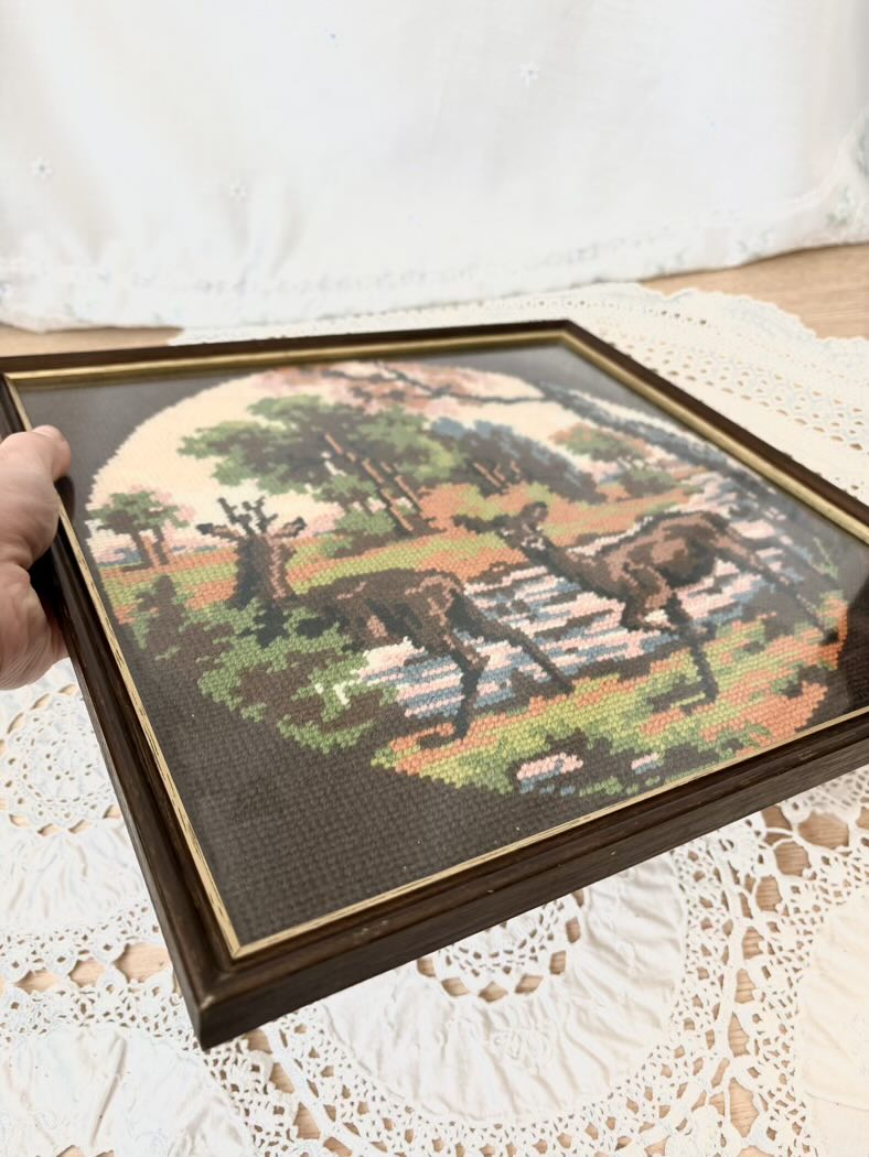Framed cross-stitched picture of deer in a forest on a lace tablecloth.