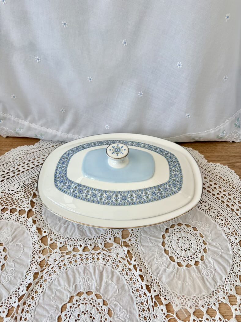 Decorative ceramic lidded container on a lace tablecloth with a white curtain in the background