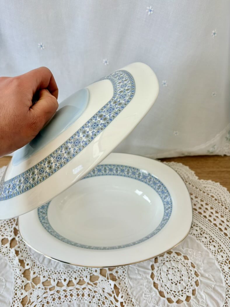 Hand holding a white plate with blue floral rim on a lace tablecloth.