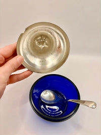 Hand holding a silver lid over a blue bowl with a spoon inside on a white background