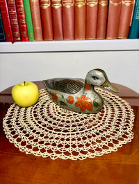 Decorative duck figurine with floral patterns on a doily, apple, and books in the background.