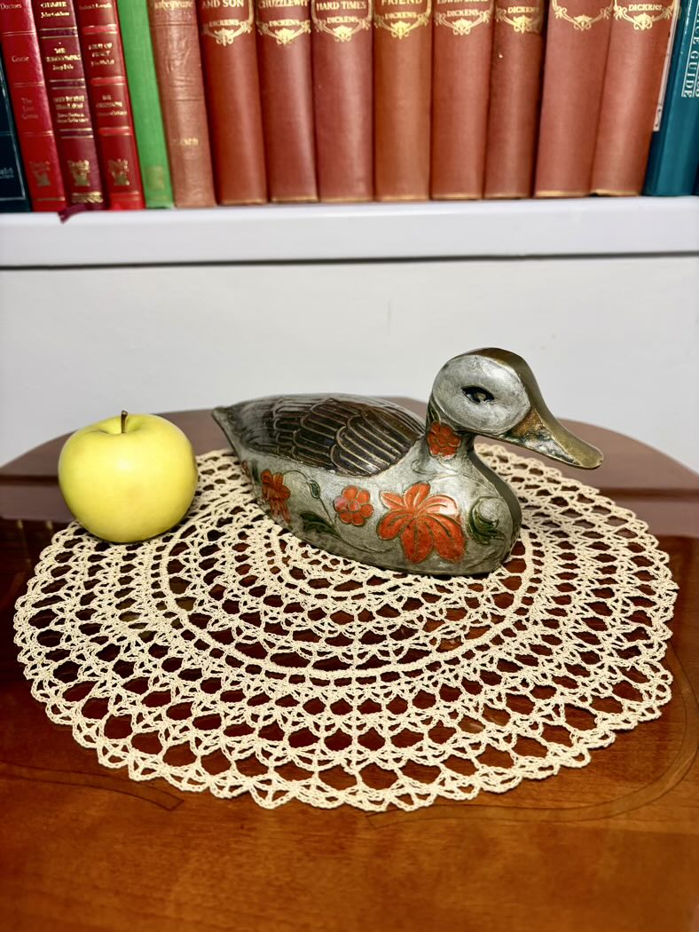 Decorative duck figurine with floral patterns on a doily, apple, and books in the background.