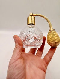 Clear glass perfume bottle with gold spray nozzle held in a hand against a neutral background