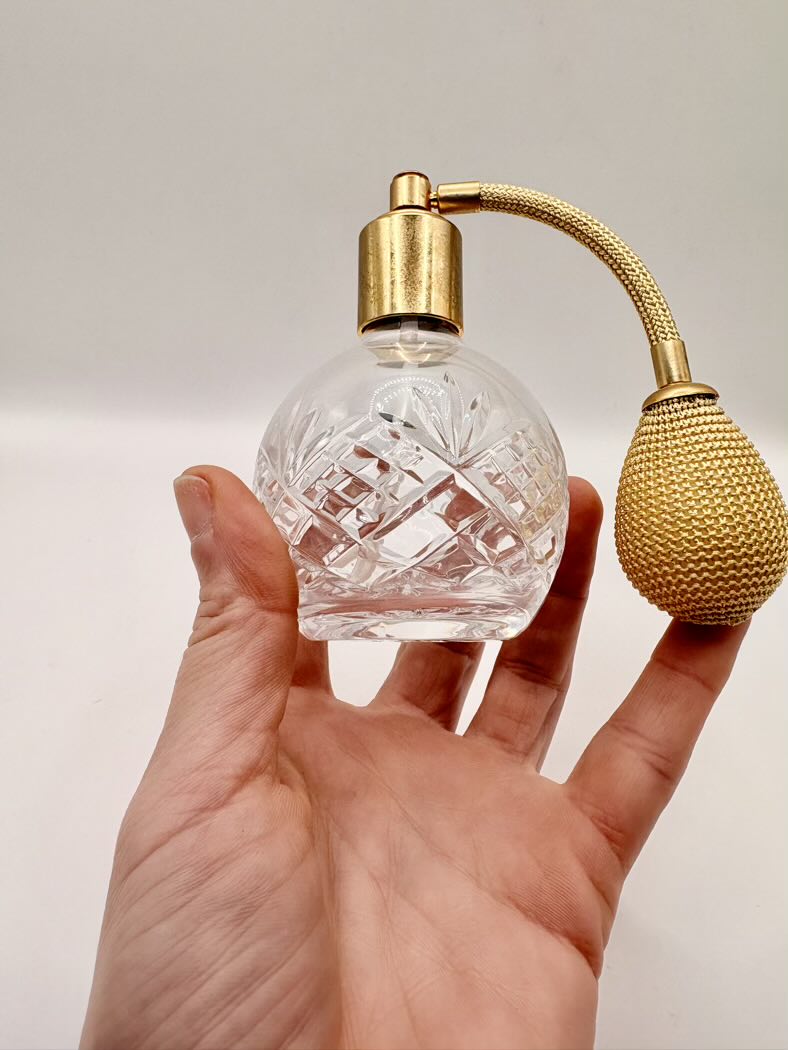 Clear glass perfume bottle with gold spray nozzle held in a hand against a neutral background