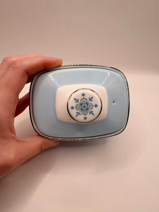 Hand holding a small ceramic container with decorative lid on a beige background