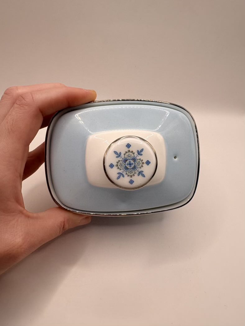 Hand holding a small ceramic container with decorative lid on a beige background