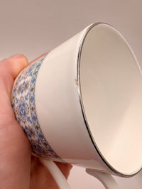 White ceramic cup with floral patterned rim held by a hand against a neutral background