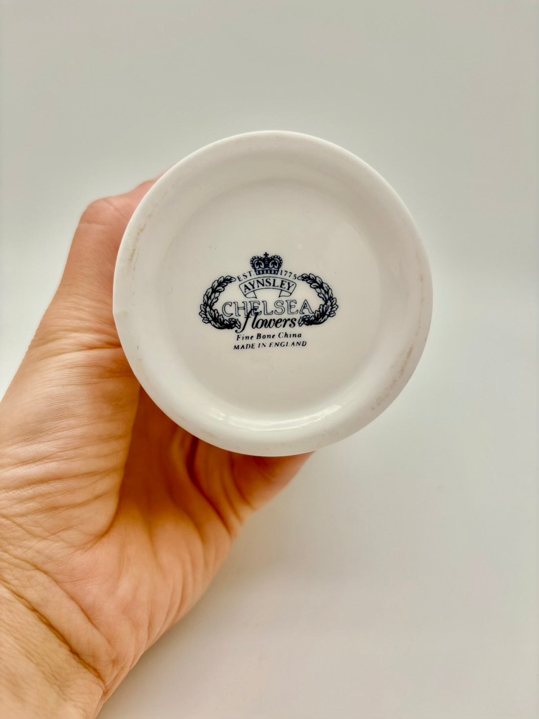 Hand holding a small white ceramic dish with a logo and text on a plain background