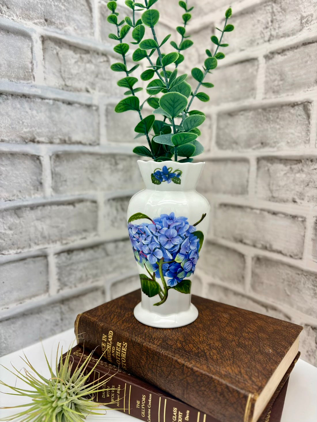 Decorative vase with blue flowers on a stack of books against a brick wall.