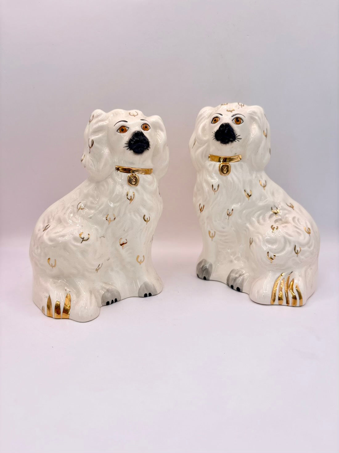 A pair of white ceramic decorative dogs with gold accents, black noses, and gold collars, set against a plain light gray background.