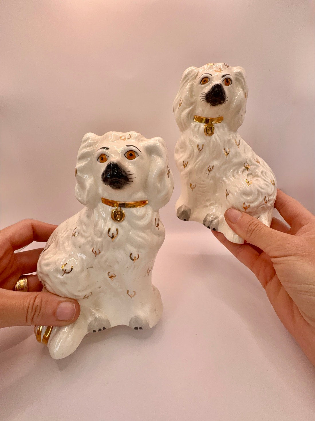 Two white ceramic dog figurines with gold accents and black noses, held up by two hands against a plain light background.