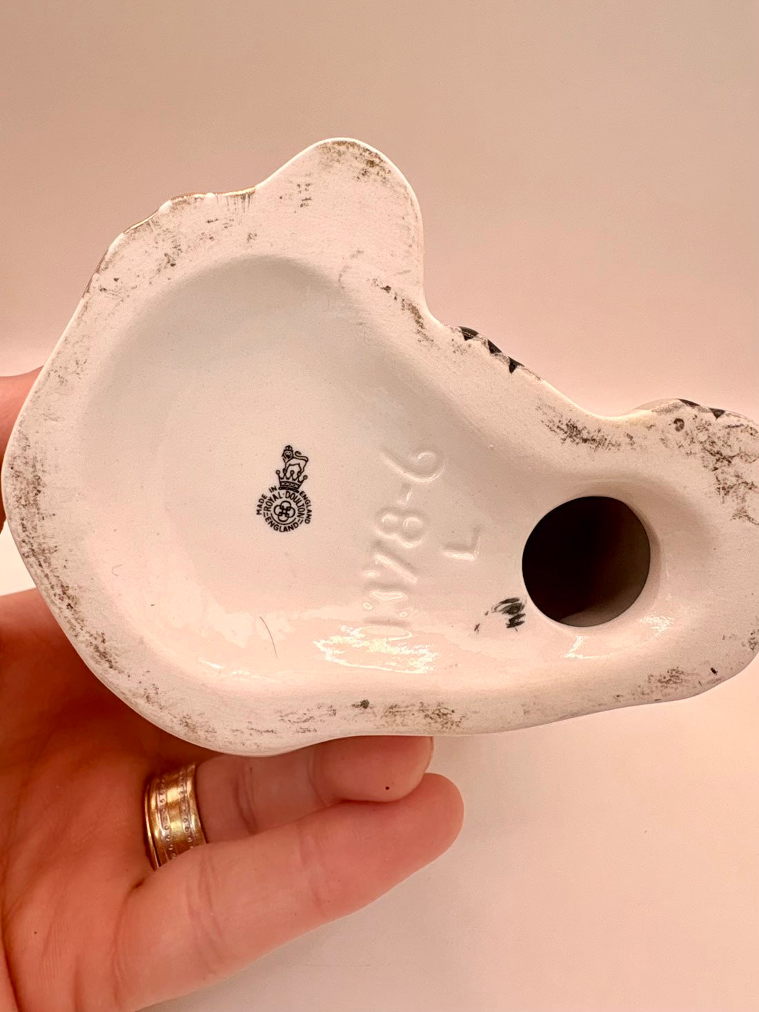 A hand holding the underside of a white ceramic object, showing a round hole and dark stamped markings on a plain light background.
