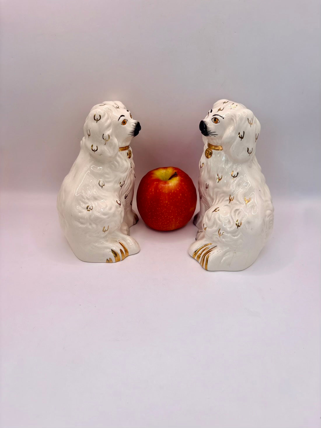 A pair of white ceramic dog figurines with gold and black detailing, positioned on either side of a red apple on a plain light background.