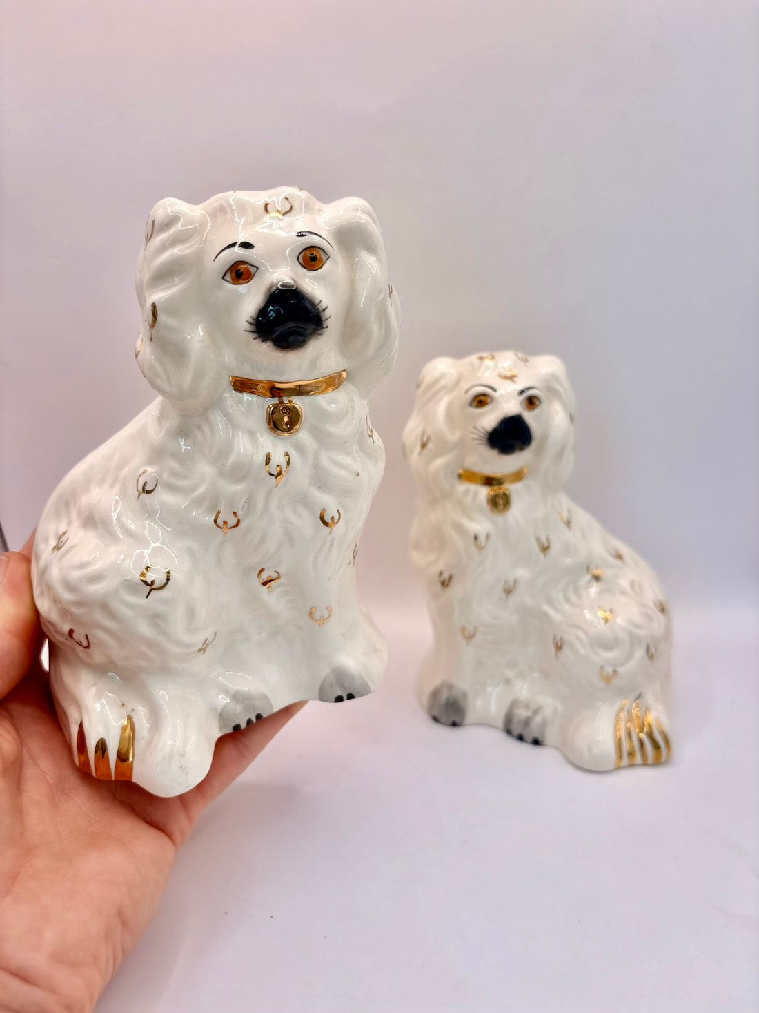Two white porcelain dog figurines with gold accents, one held in a hand and one standing on a white surface against a light background.