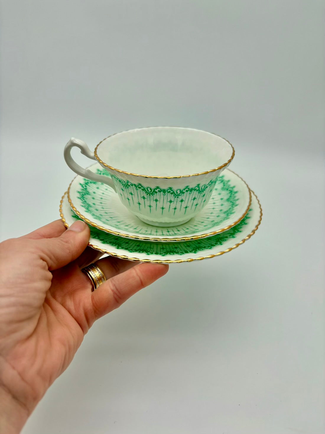 Hand holding a vintage teacup and saucer with green patterns on a plain background