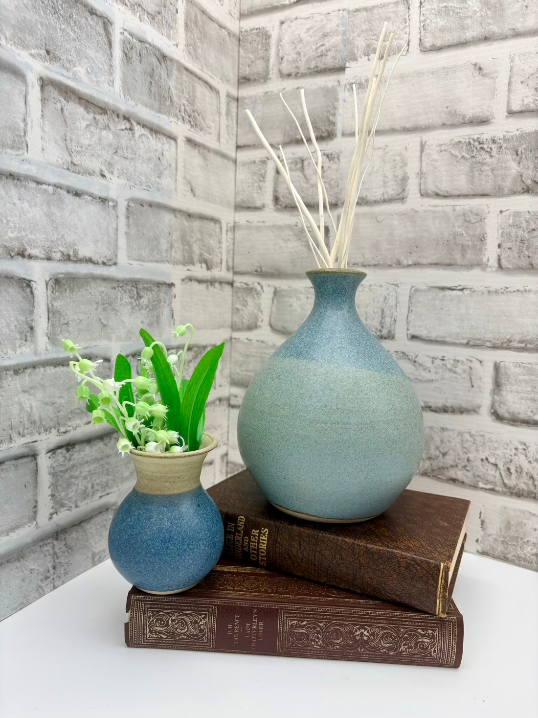 Two blue vases and a small plant on books against a brick wall.