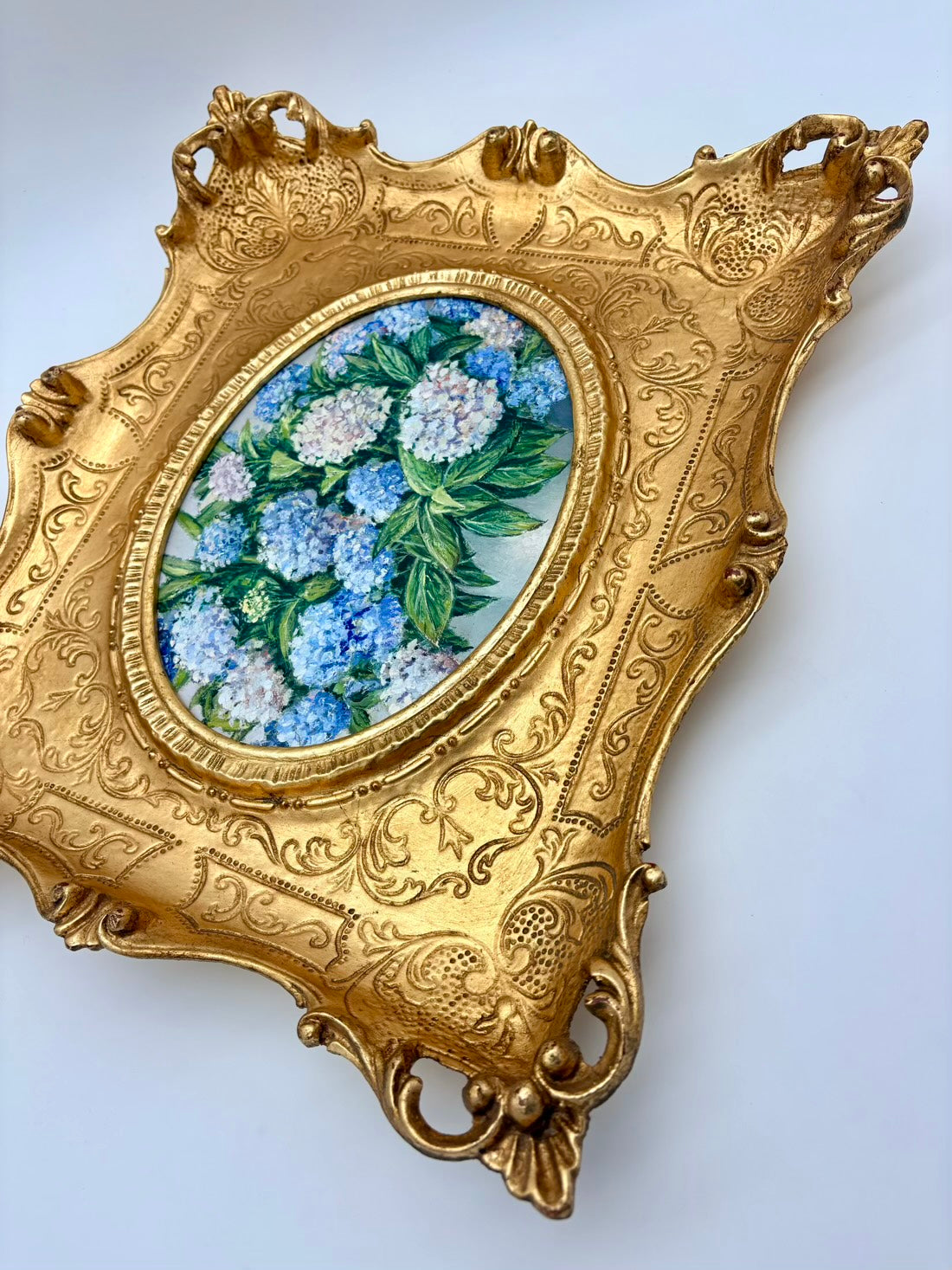 An ornate gold-colored decorative tray or wall plaque featuring an oval inset painted with blue and white hydrangeas, set against a plain light background.